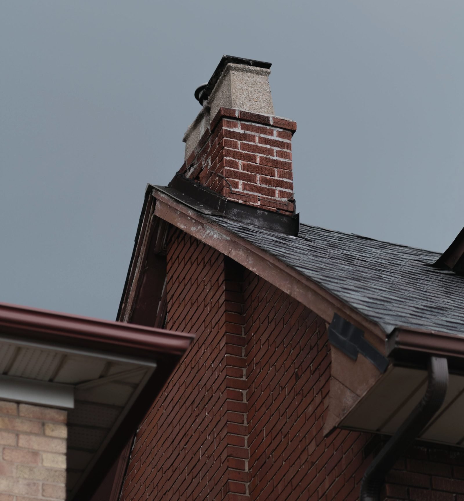 The corner of a brick house showing shingle courses, gutter, and downspout under a gray sky