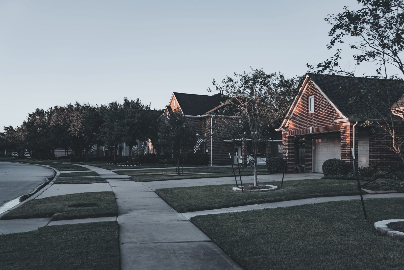 A quiet street of brick homes at dusk, an American flag on one porch